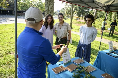 several people are standing around a table in a park like area while a person demonstrates how plant presses work according to a sign on the table between them next to small books of some kind