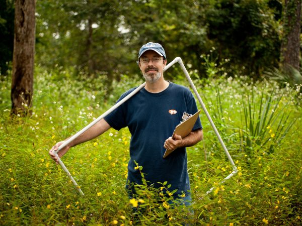 a person holding a clip board and a large square frame made from pvc pipes standing in a wooded field