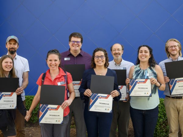 Members of the Daniels lab pose against a blue background, holding their Champions for Change award certificates