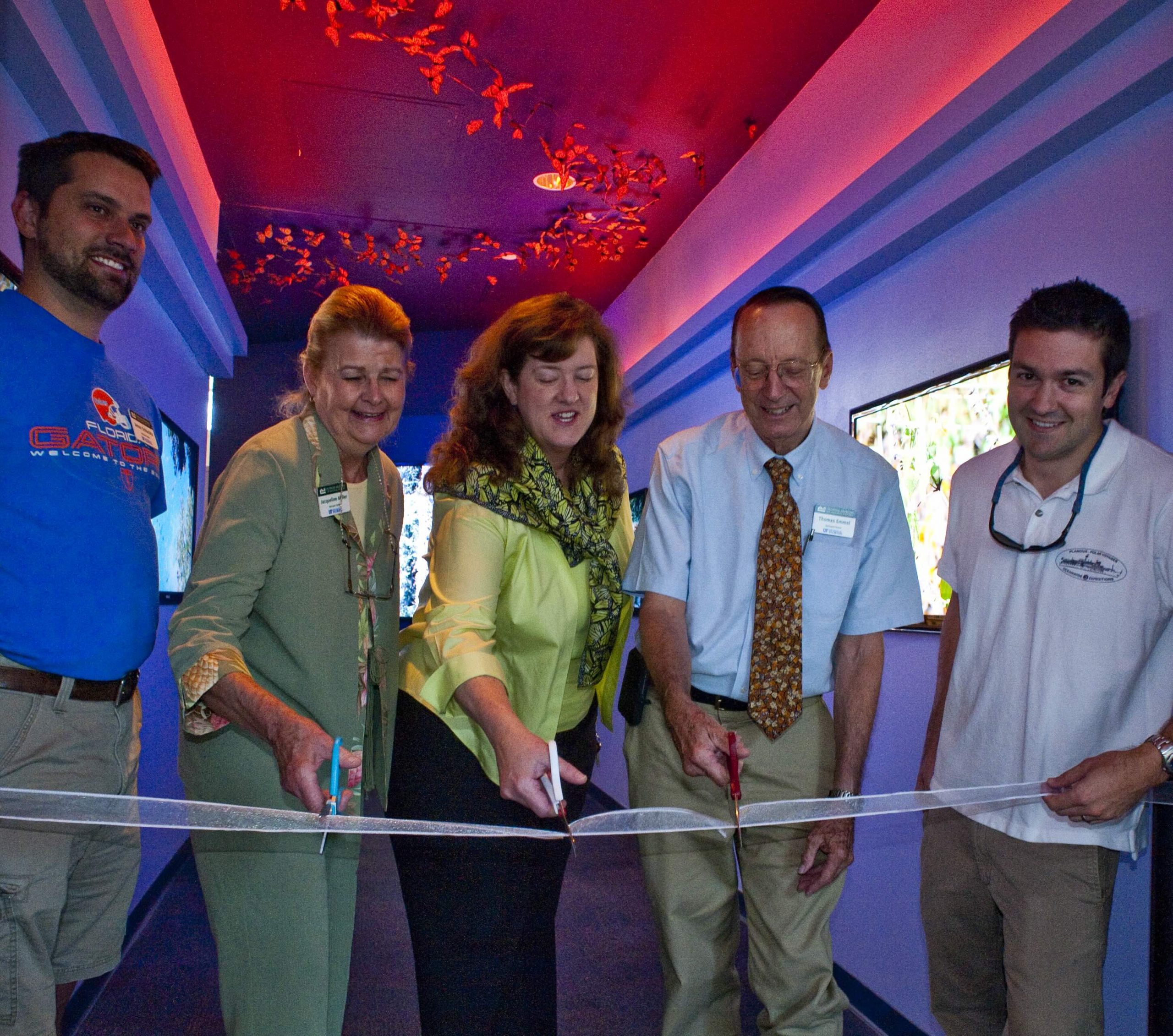 Five people side by side. The two at opposite ends of the line are holding a ribbon, while the three in the middle use scissors to cut it. 