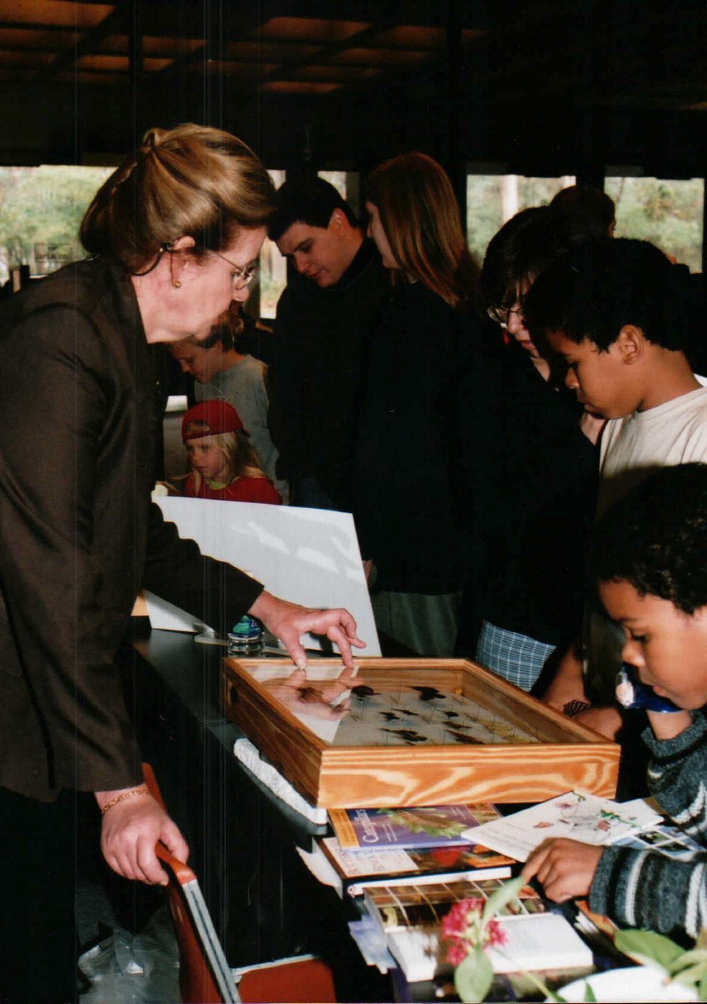 Person behind a table shows butterflies in a display case to people on the opposite side.