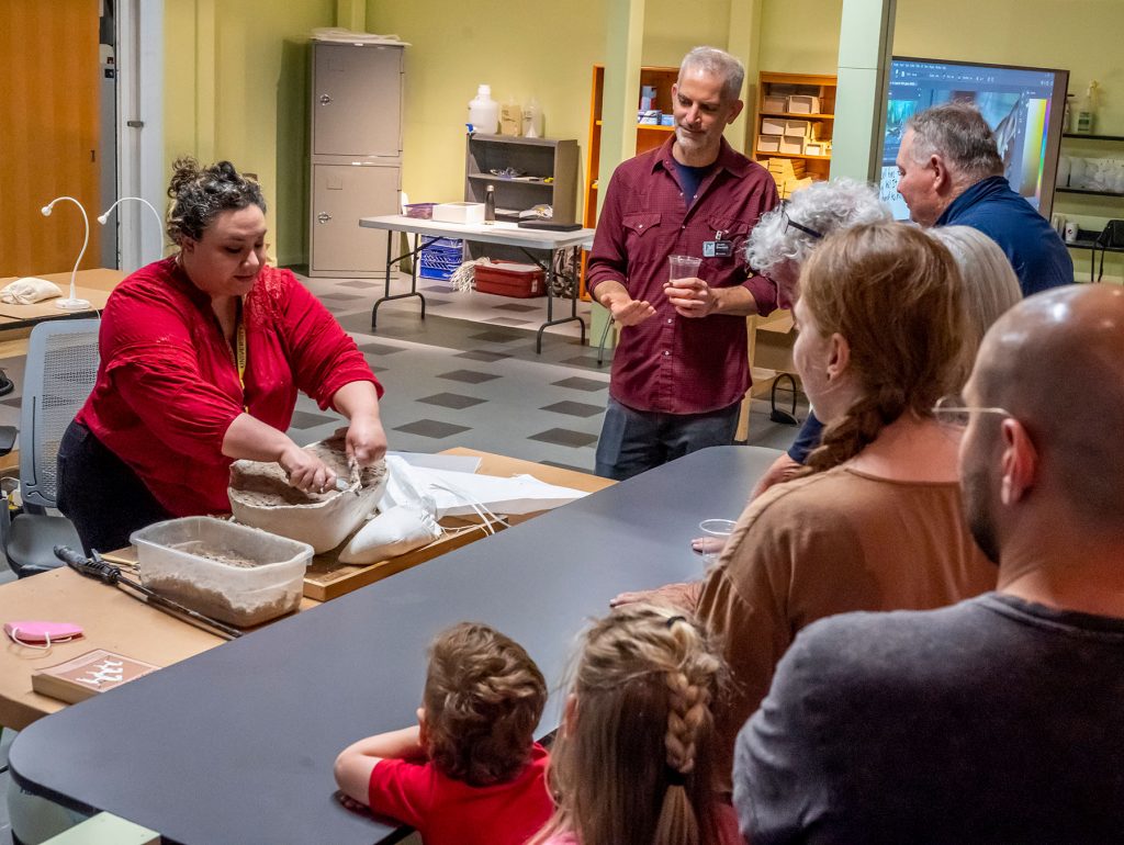 Person tears away at the plaster surrounding a fossil while visitors watch.