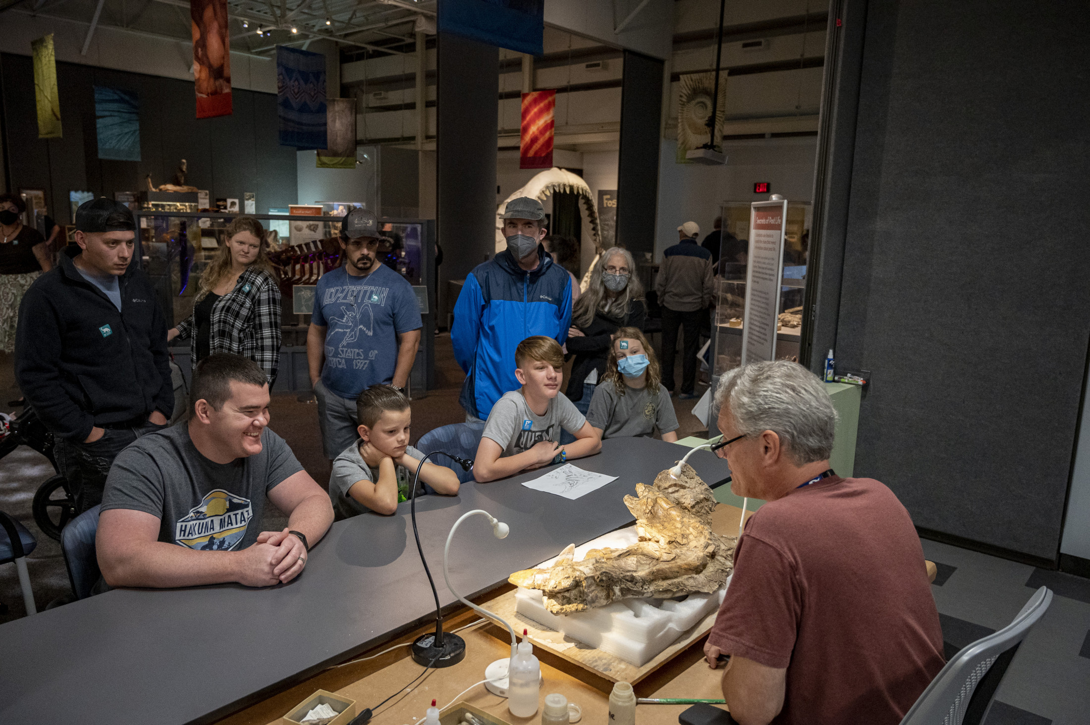 Person working at a desk with a large fossil on it talks with a crowd of visitors at a museum exhibit. 