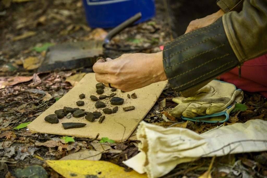 Close up of pottery fragments sitting on a board on the forest floor as someone picks up a piece to examine it.