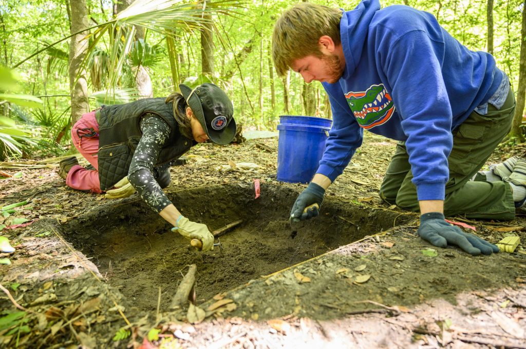 Two students using hand shovels to dig an archaeology pit.