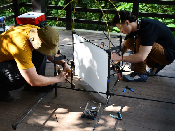 Two people assemble a biodiversity monitoring device on a deck in a rainforest.