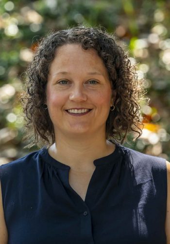 White woman with brown curly hair wearing navy blue top in front of green foliage background