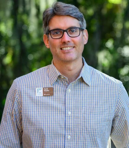 White man wearing glasses and checkered shirt standing in front of green foliage