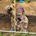 person kneeling in an excavation unit to measure a stratigraphic profile