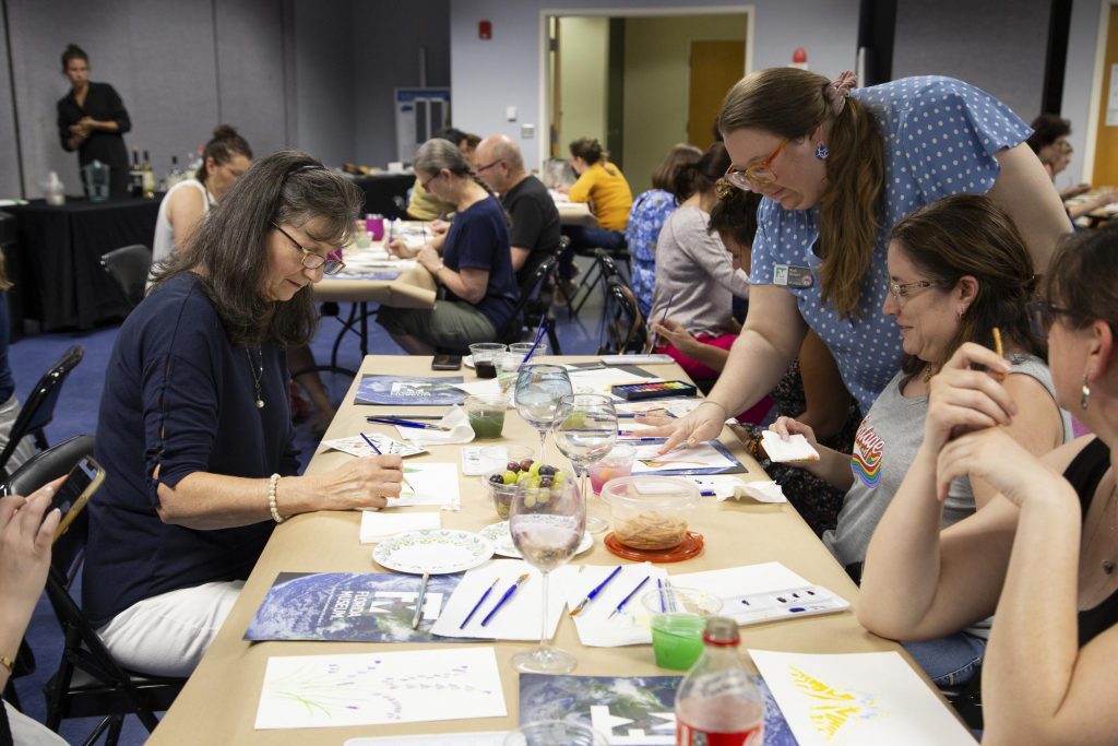 Watercolor and wine participants sit around a table working on their artwork and speaking with an event assistant