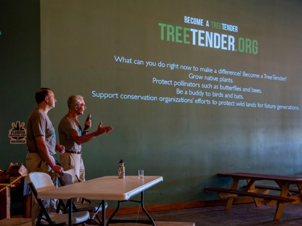 two people stand behind a white folding tab, one is speaking into a microphone, information is projected onto the wall behind him