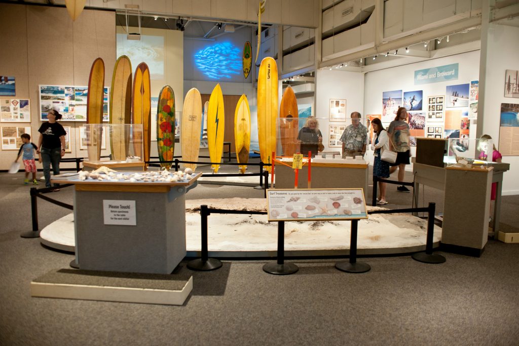 visitors in the surfing Florida exhibit look at various displays. In the center of the room is a line of surfboards in various shapes and colors