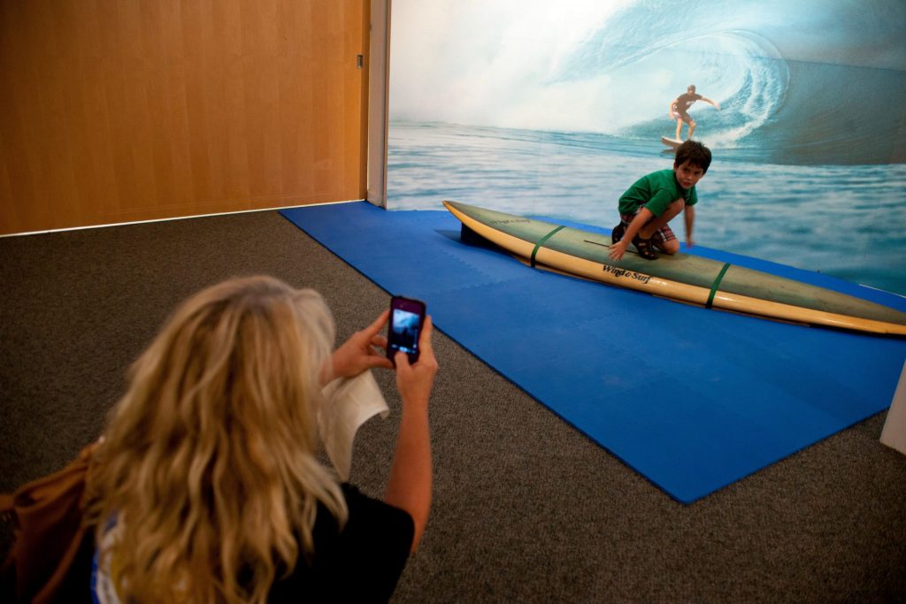 person takes a cell phone photo of a child on a display surfboard set in front of a large photo of a curling wave