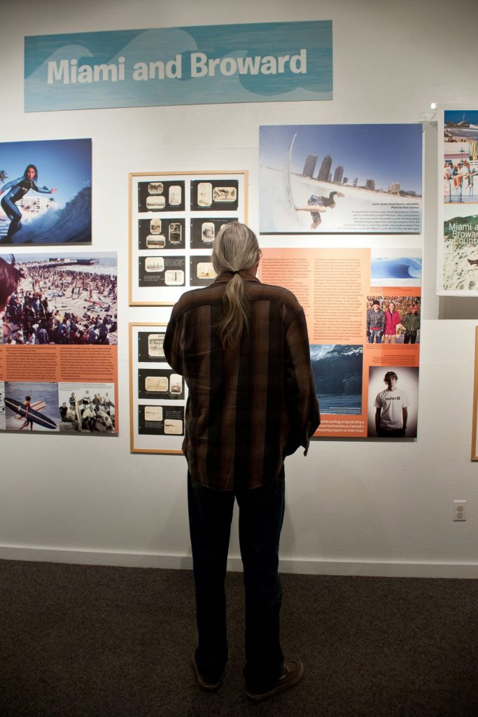 person with long hair stand in front of a wall filled with color and black and white photos of people surfing. All the images are under a sign that reads Miami and Broward