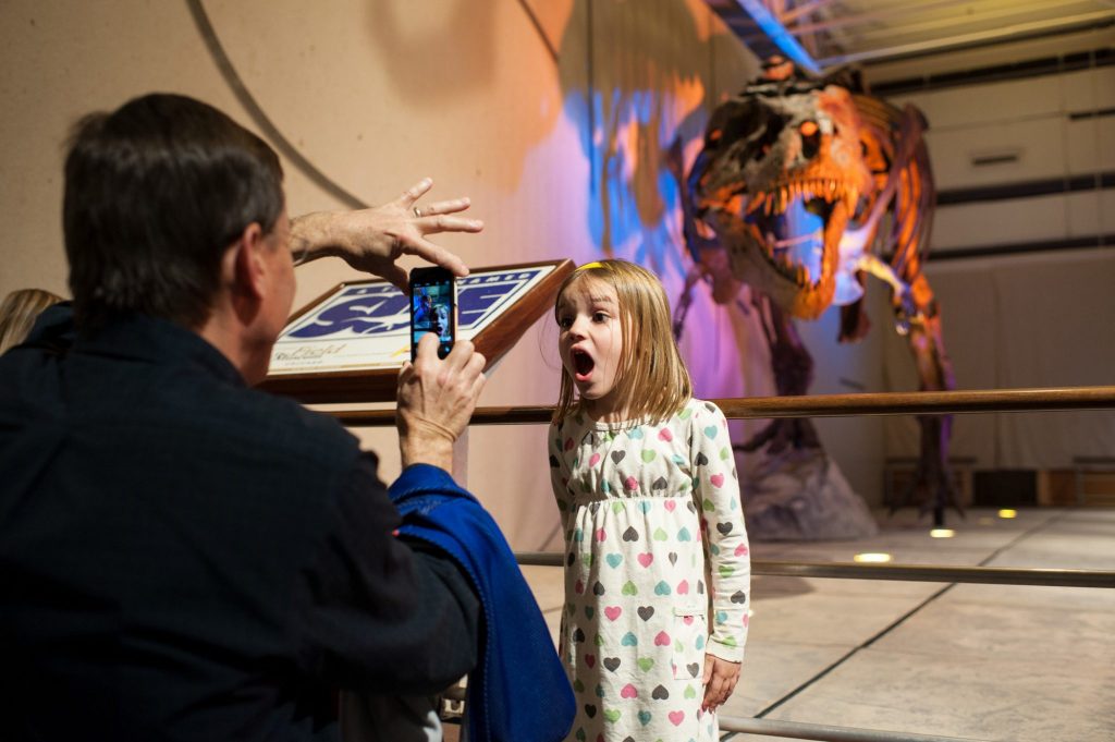 young girl being photographed standing in front of Sue, the fully articulated, life-size t-rex skeleton
