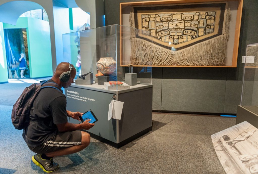 visitor wearing head phones and holding a table kneels next to a display of pottery and listens to the audio portion of each exhibit