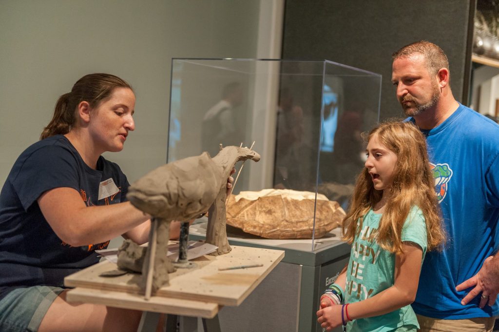 young visitor speaks with researcher and artist who is working with clay to create a model