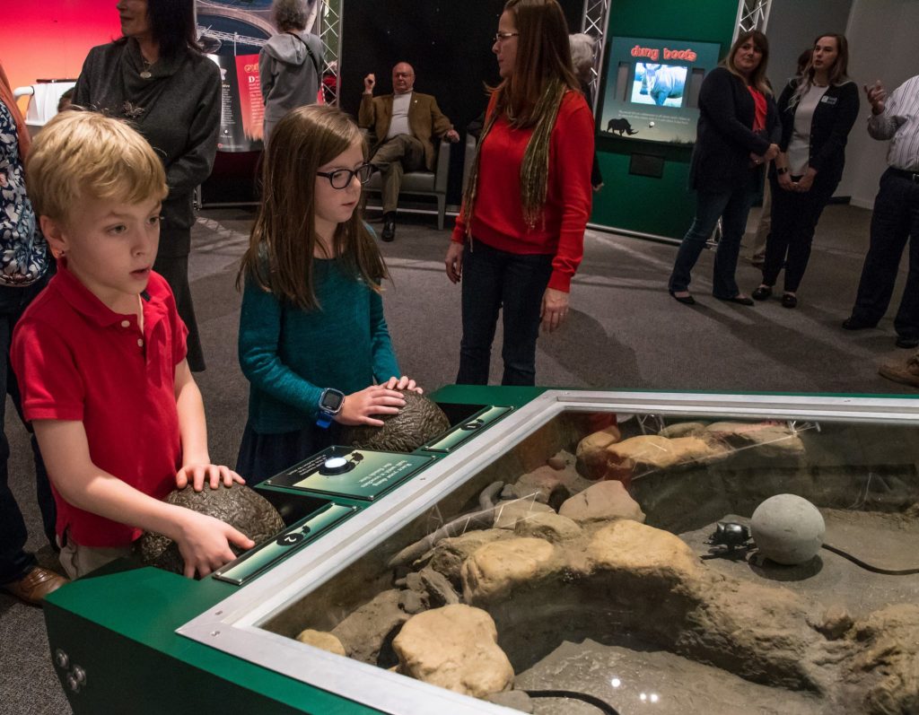 two children using iterative exhibit display behind them many people explore the exhibit