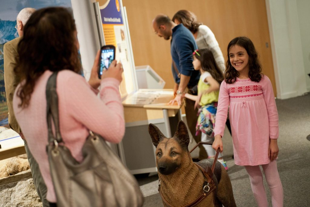 child holds the harness of a fabricated seeing eye dog and smiles as an adult takes a photo with a camera phone