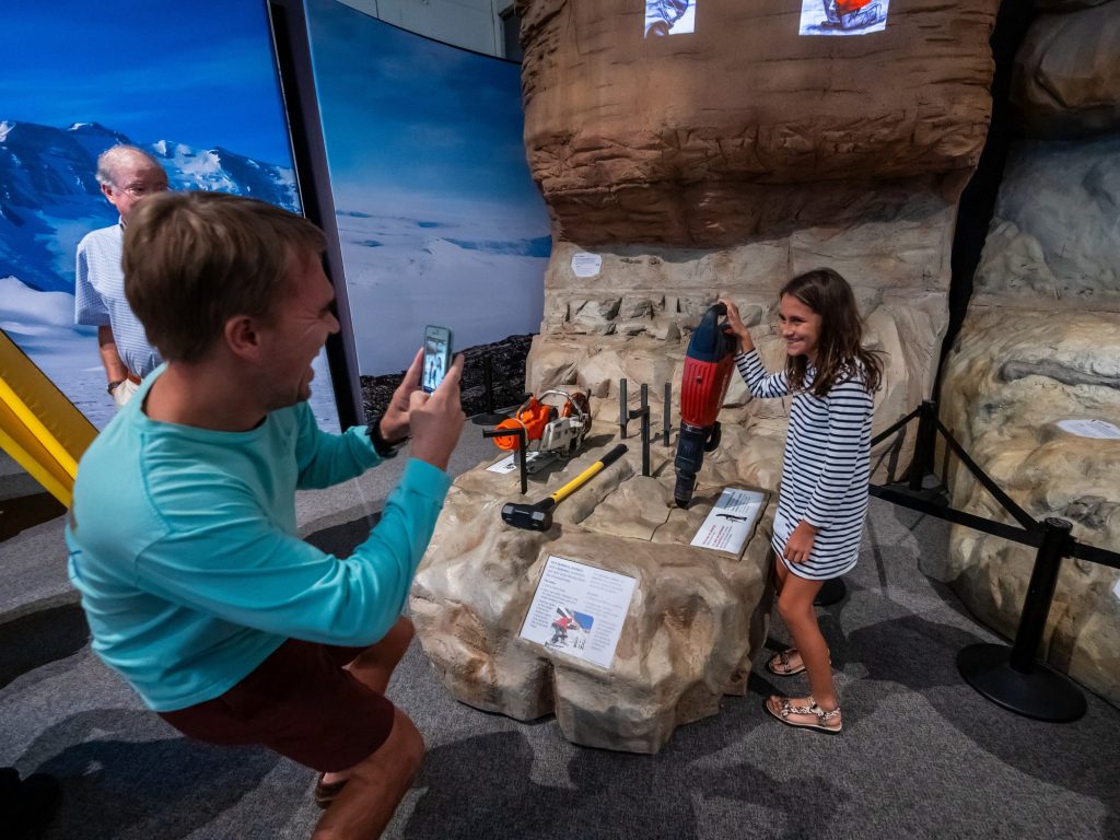 adult photographing a child standing next to the digging tools on display in the Antarctic Dinosaurs exhibit