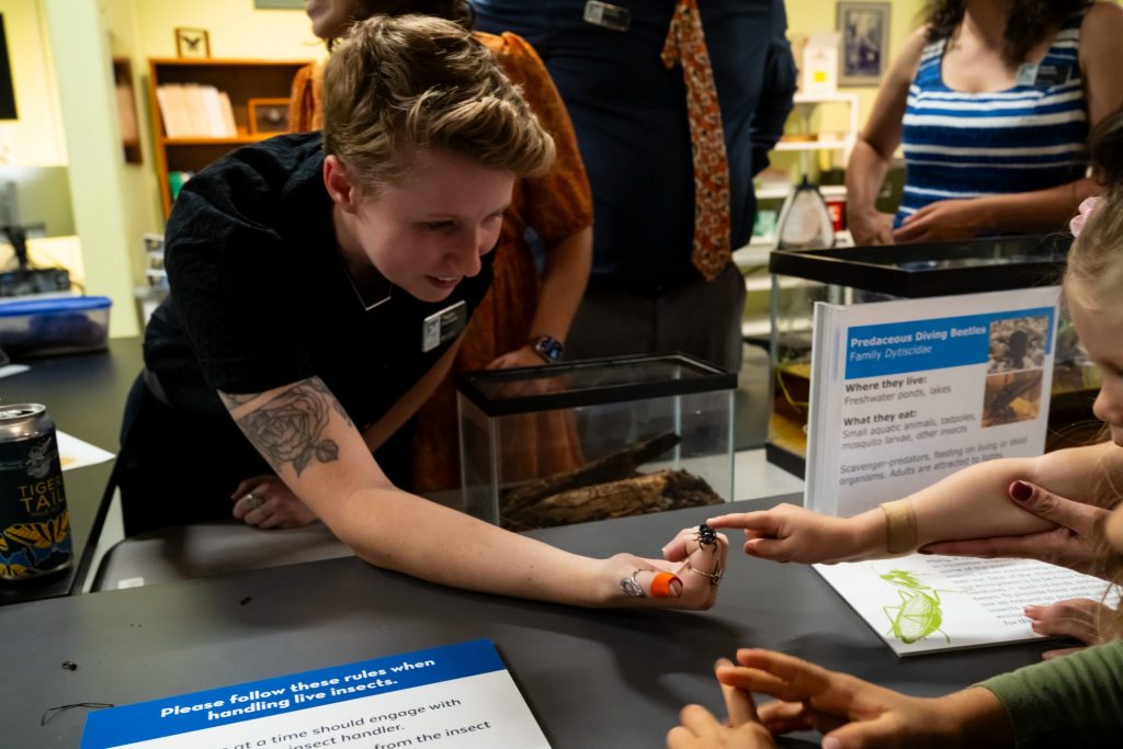 Florida Museum Researcher leans over a table to allow a young child to touch the beetle in her hand