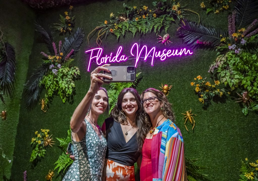 three people are taking a selfie in front of a wall covered in fake leaves and insects and a neon sign that reads Florida Museum