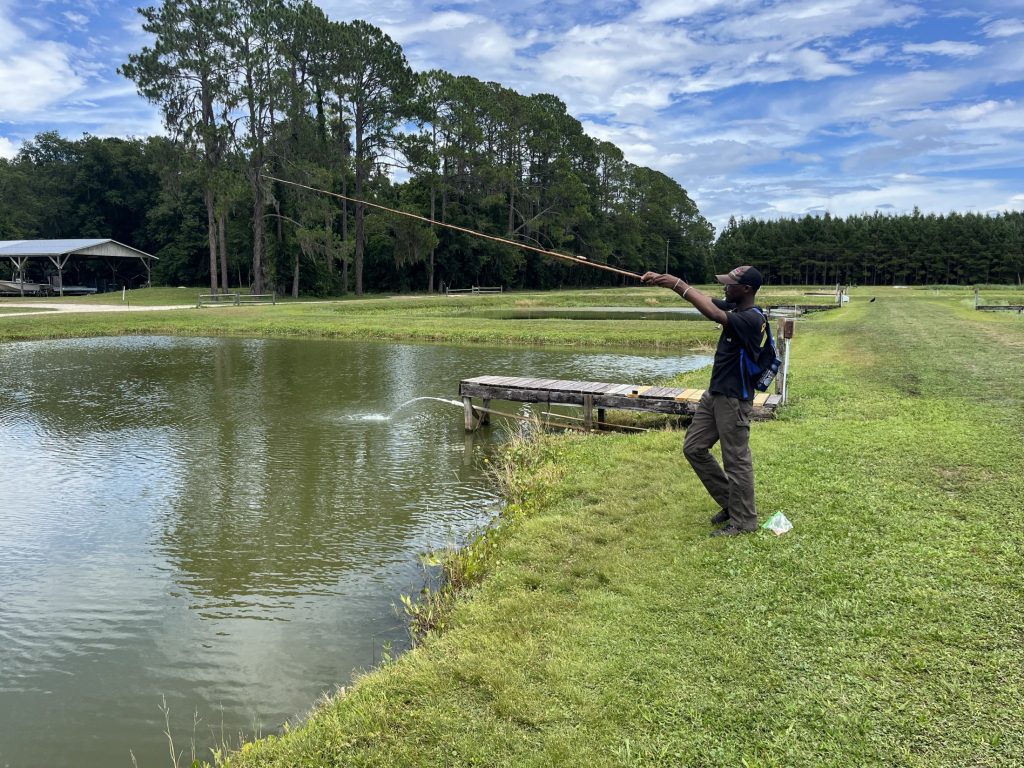 Wide shot of Joel Saunders throwing out a fishing line into a pond. with trees in the horizon in the background.