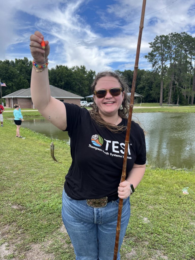 Laura smiles and holds up the fish she caught in one hand.