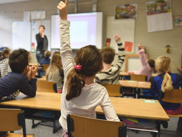 Student-view of a classroom. Students are raising their hands in front of a smart board.